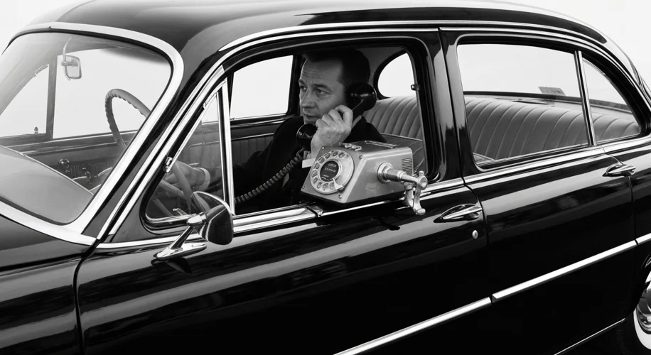 A vintage black and white photograph of a person using an early car phone from the 1950s. The car is